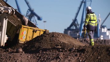 Agence Satellite – tournage vidéo sur le chantier du quai H au port de Sète avec un ouvrier en sécurité observant un camion déversant des matériaux, fond de grues portuaires.